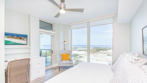 Une chambre avec un grand lit, un fauteuil avec un coussin jaune, un téléviseur fixé au mur et une vue sur la plage grâce à de grandes fenêtres.