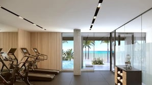 A modern gym with exercise equipment, a glass wall offering a view of a pool and palm trees, and a small kitchenette area. - Luxurious Oceanfront Condo with Rooftop Pool on Brighton Beach– Sleeps 4 (Bridgetown)