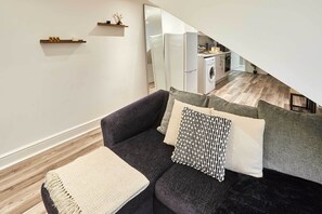 Sala moderna com um sofá cinza escuro, uma mesinha de centro e um espelho grande que reflete a área da cozinha.