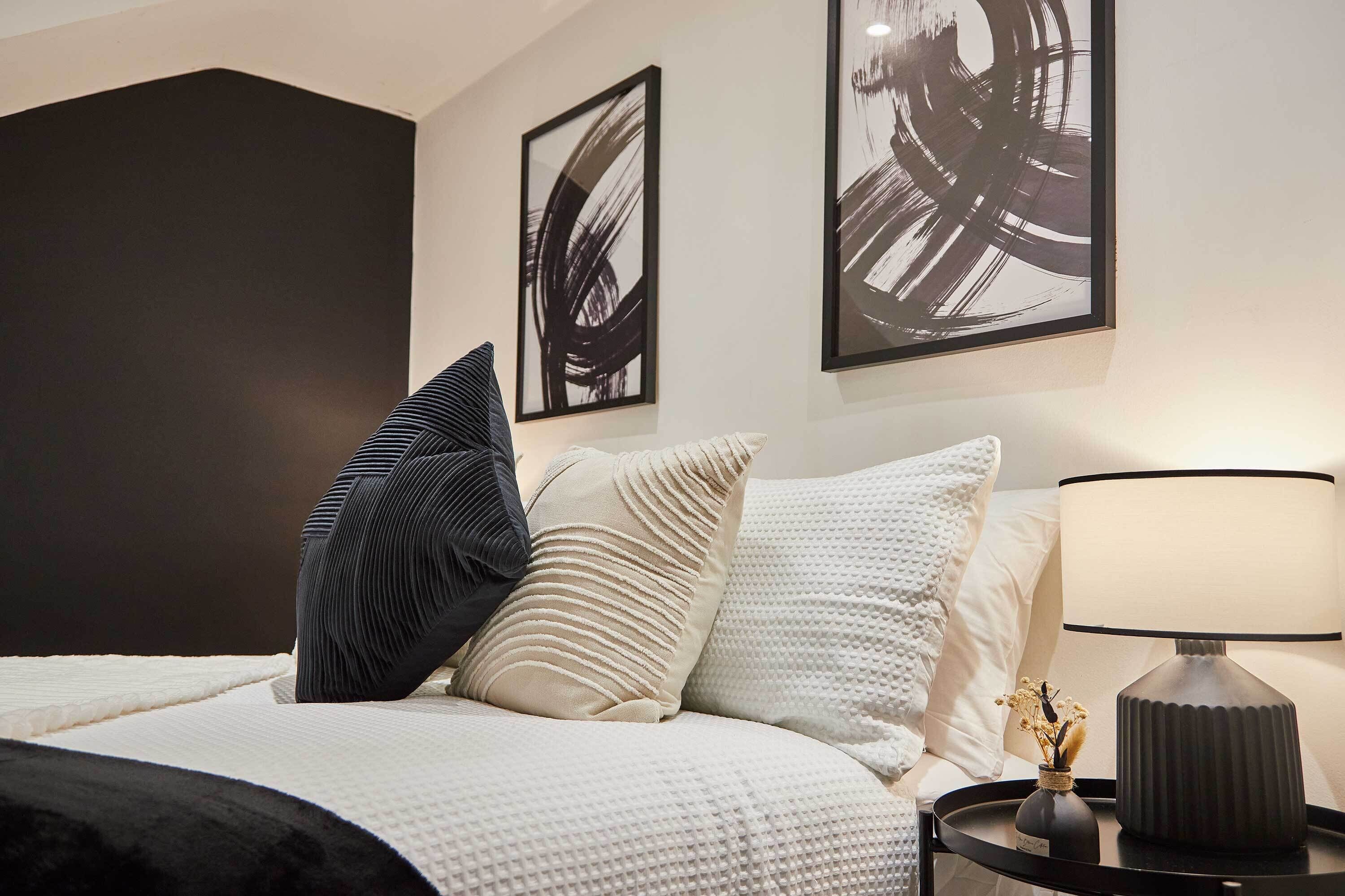 A neatly made bed with a white and black striped pillow, a beige pillow, and a textured white pillow. There is a black lamp on a bedside table and two abstract paintings on the wall.