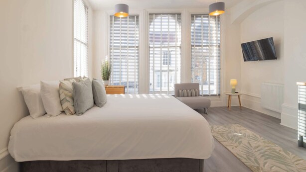A bedroom with a large bed, two pendant lights, a chair, a small table with a lamp, and a view through large windows. - Host & Stay | Harbour Rest (Ramsgate)