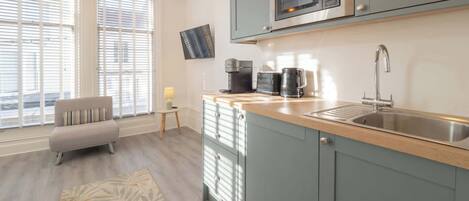 Une cuisine moderne équipée d’un four à micro-ondes encastré, d’une machine à café et d’un évier. Le salon adjacent comprend un canapé et une petite table avec une lampe.