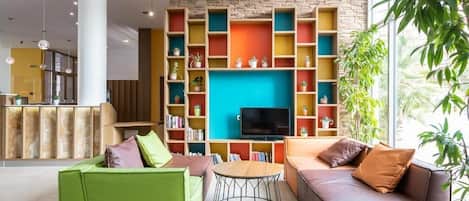 Una sala de estar moderna con un televisor grande, estanterías coloridas, un sofá verde, un sofá marrón y una mesa de centro redonda.