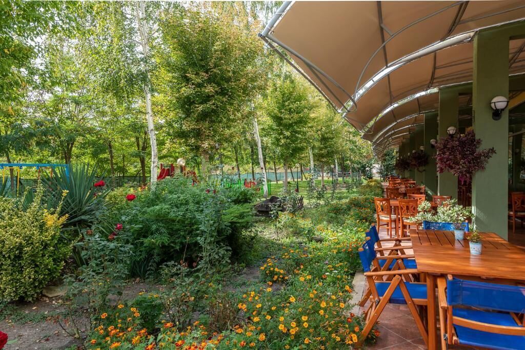 An outdoor dining area with wooden tables and chairs, surrounded by lush greenery and flowering plants.