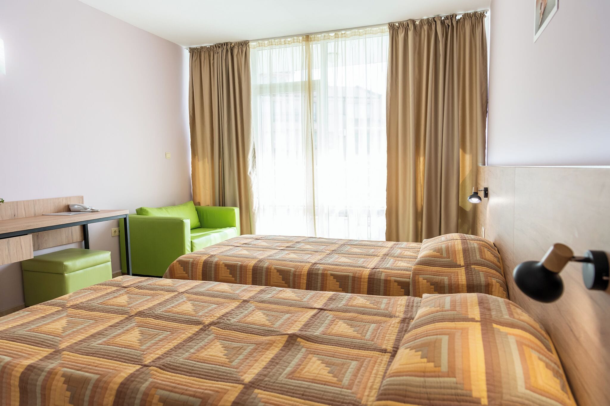 Ein Hotelzimmer mit zwei Betten, einem Schreibtisch, einem grünen Stuhl und einem Fenster mit Vorhängen.