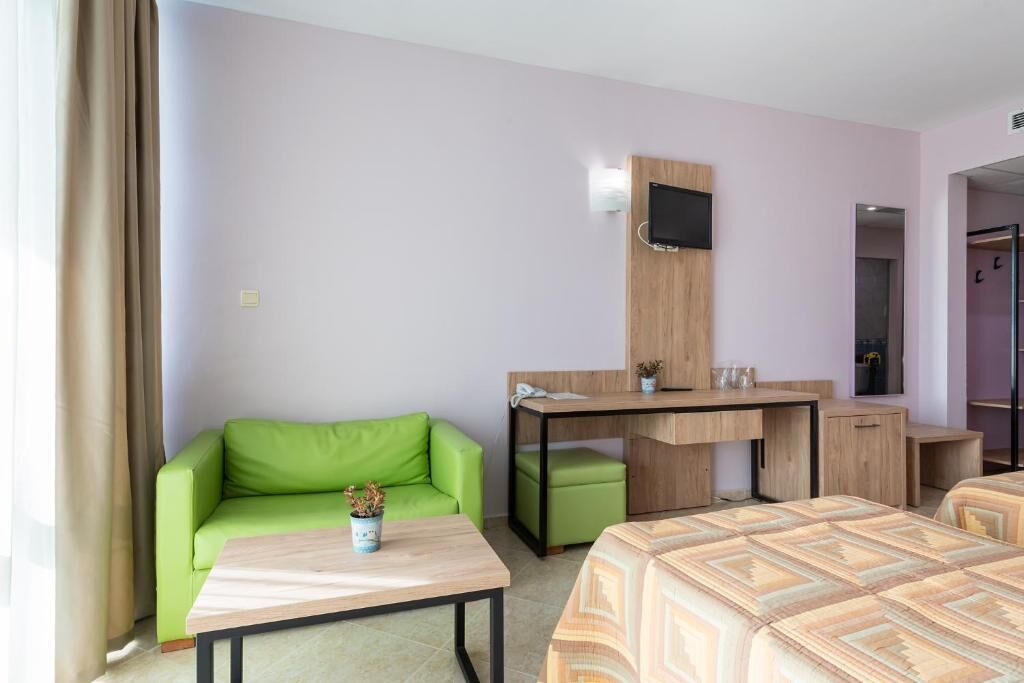 A hotel room with a green sofa, a wooden desk, a bed with patterned bedding, and a TV mounted on the wall.
