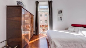 A hotel room with a wooden wardrobe, a bed with white bedding, a red headboard, and a balcony with a view of buildings.