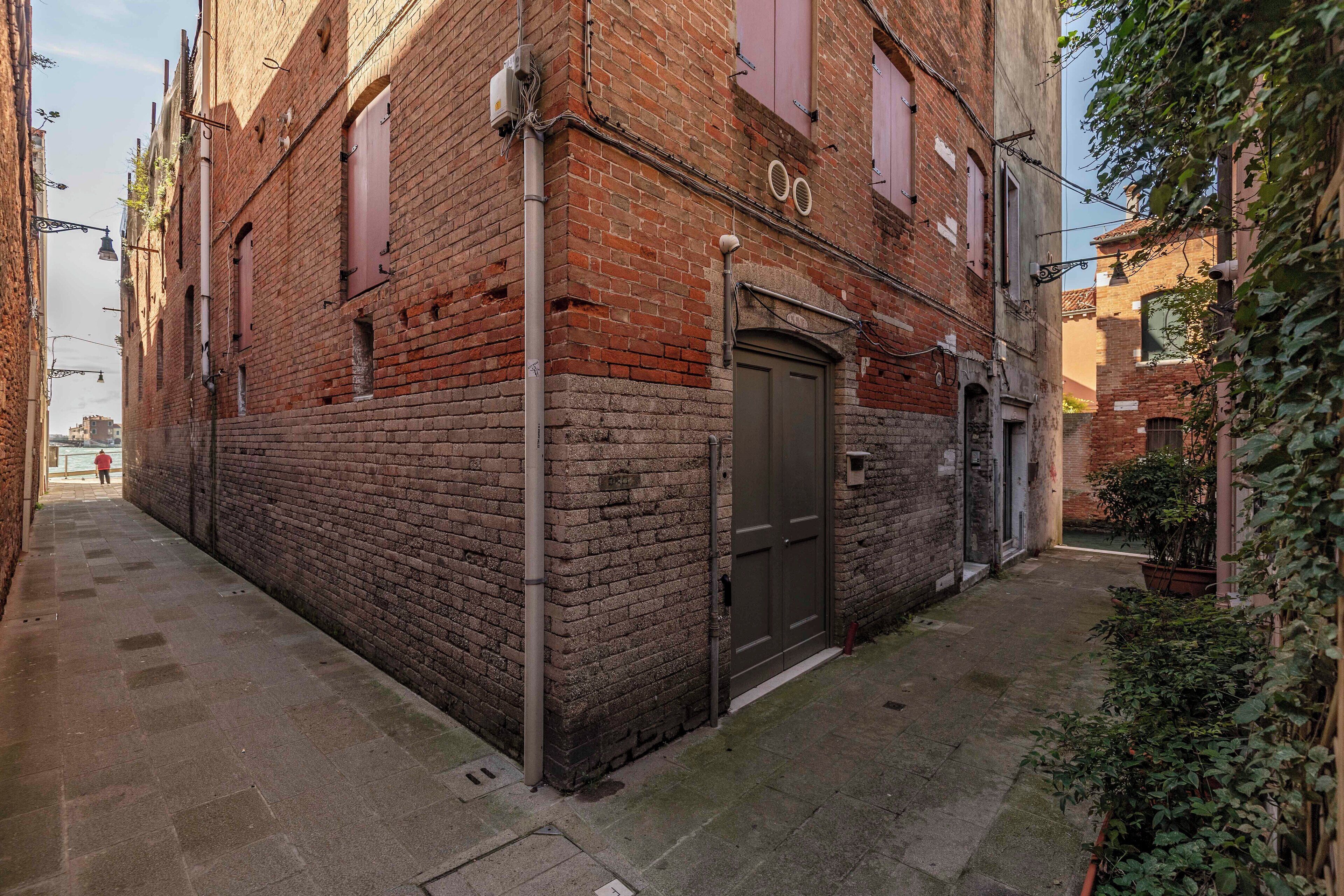 A narrow alleyway with a brick building, a closed door, and a small garden area with plants.