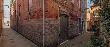 A narrow alleyway with a brick building, a closed door, and a small garden area with plants.