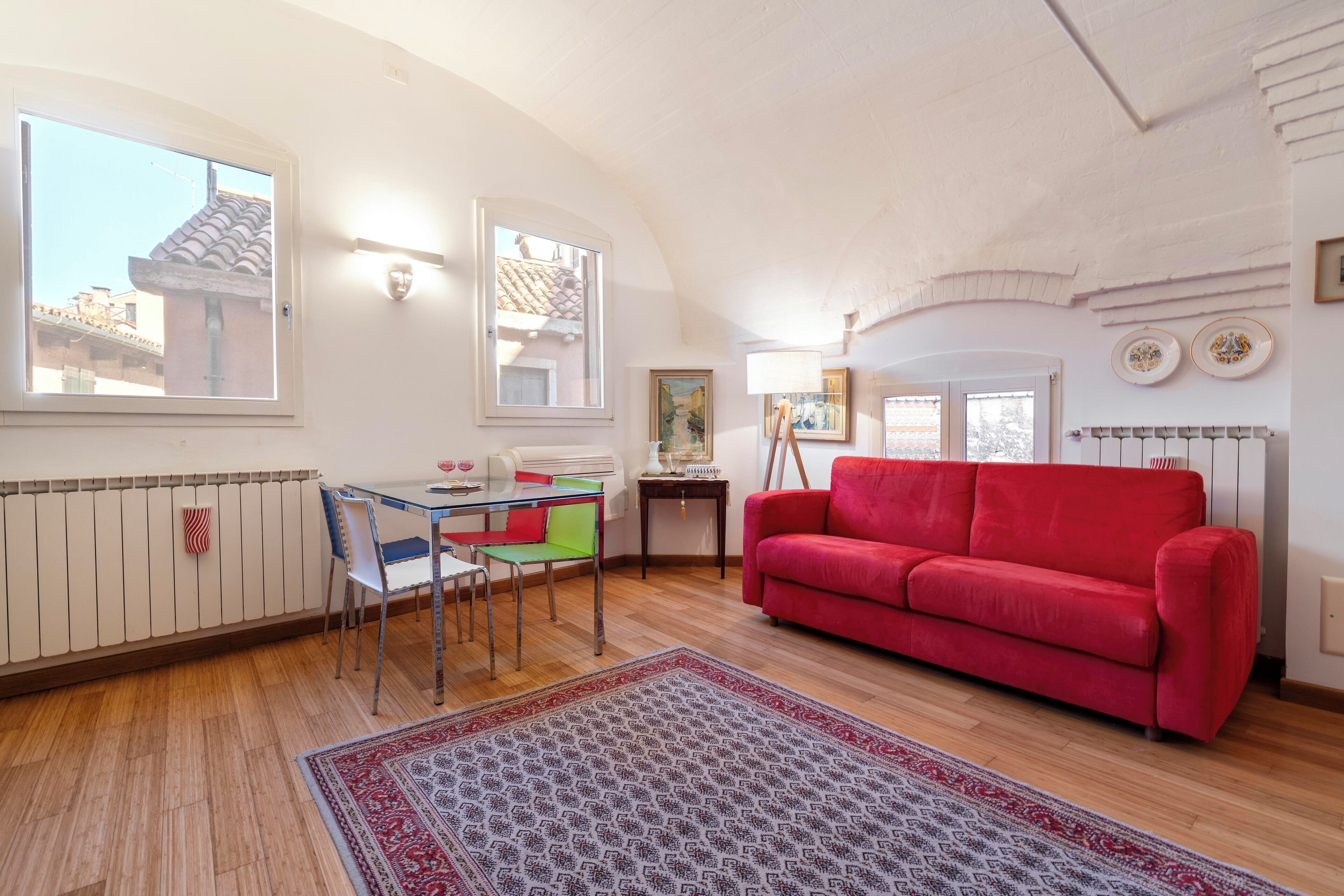 A living room with a red sofa, a dining table with chairs, a patterned rug, and a radiator.