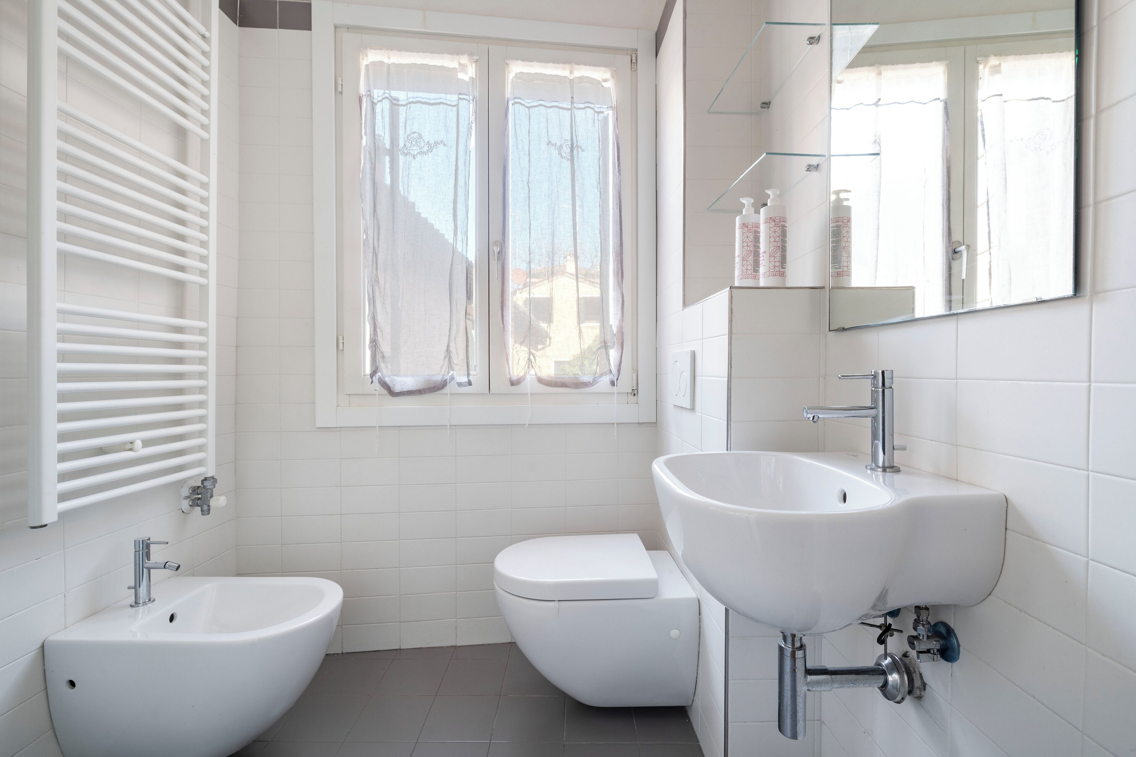 A modern bathroom with a bidet, toilet, and sink, featuring white fixtures and a window with curtains.