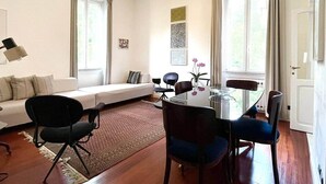 Een moderne woonkamer met een glazen eettafel, een bank en een salontafel. Er zijn twee ramen met gordijnen, een kroonluchter en een deur.