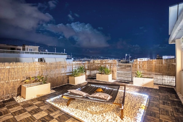 A rooftop lounge area with a bed, potted plants, and a view of the city at night.