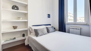 A bedroom with a bed, a window with blue curtains, a radiator, and shelves with decorative items. - Glorioso Marble | Trastevere ArtDesignApartments (Roma)