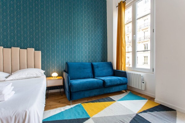Een slaapkamer met een bed, een blauwe bank, een nachtkastje met een lamp, en een raam met gordijnen.