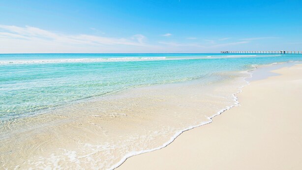 A beach with clear turquoise water, a sandy shore, and a pier extending into the distance.