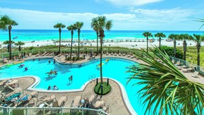 A swimming pool with lounge chairs and palm trees, overlooking a beach with umbrellas and people.