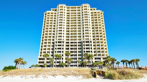 A tall, multi-story hotel with balconies, located on a beach with palm trees and sandy ground.