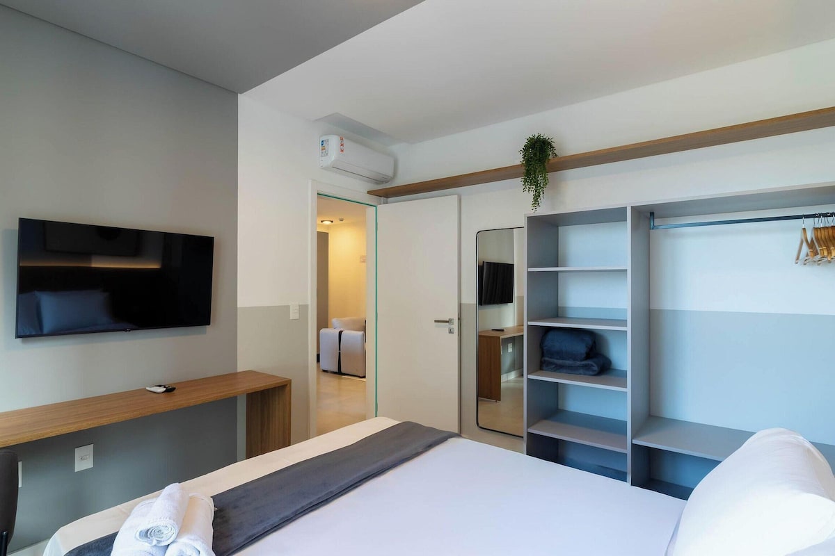 A modern hotel room with a bed, a TV mounted on the wall, a wardrobe with shelves, and a small desk with a chair.