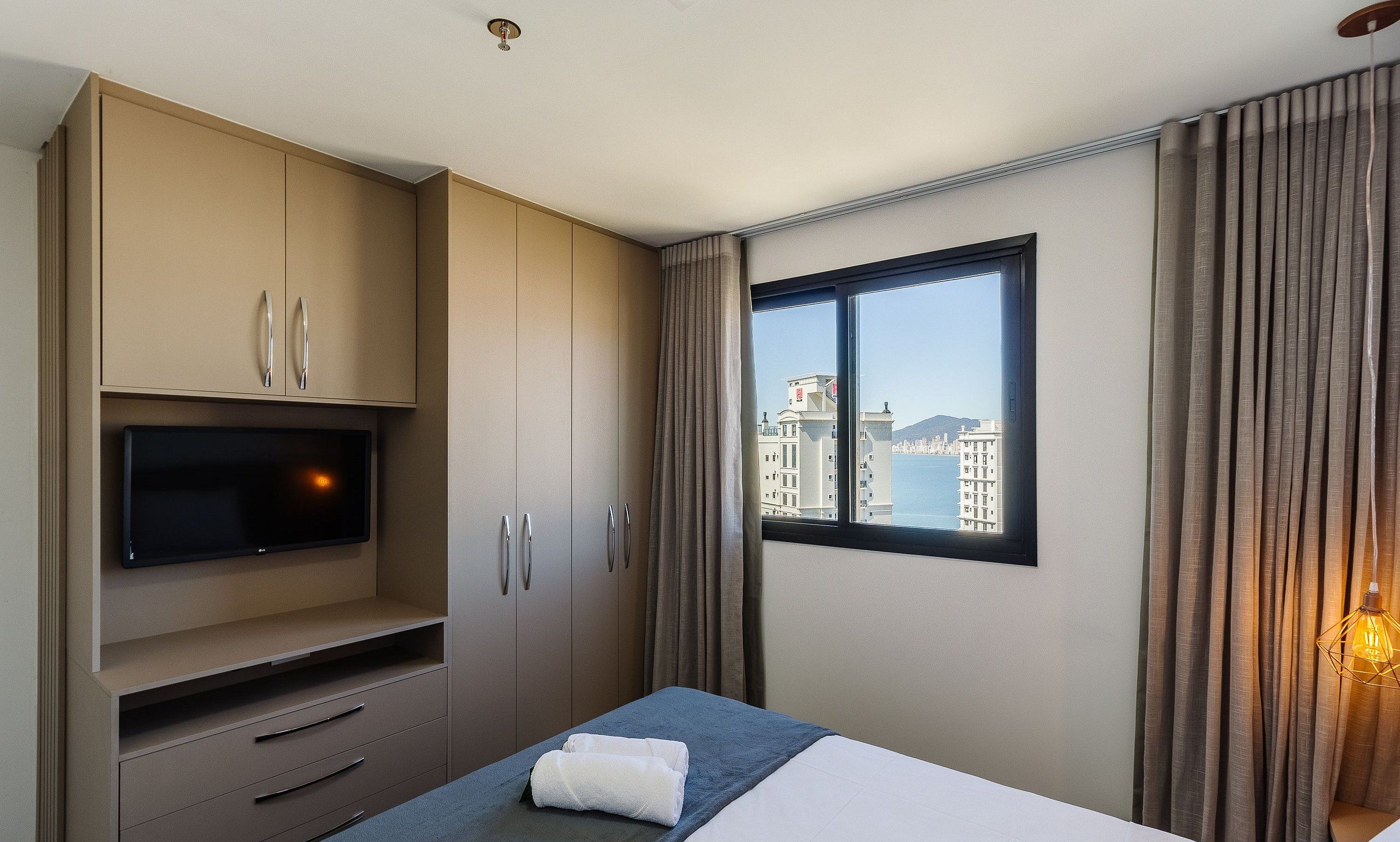 A hotel room with a bed, a TV, a wardrobe, and a window with a city view.