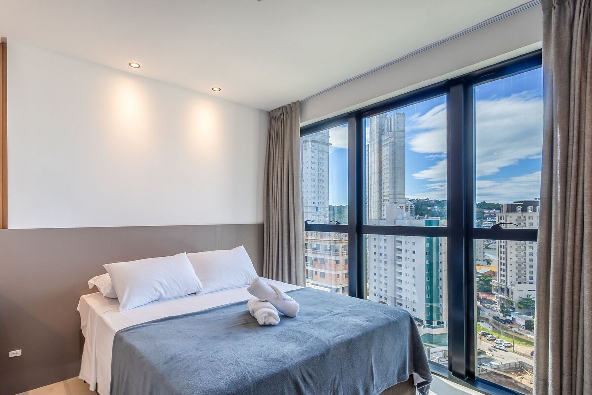 A modern hotel room with a large bed, a bedside table, and a view of the city through floor-to-ceiling windows.