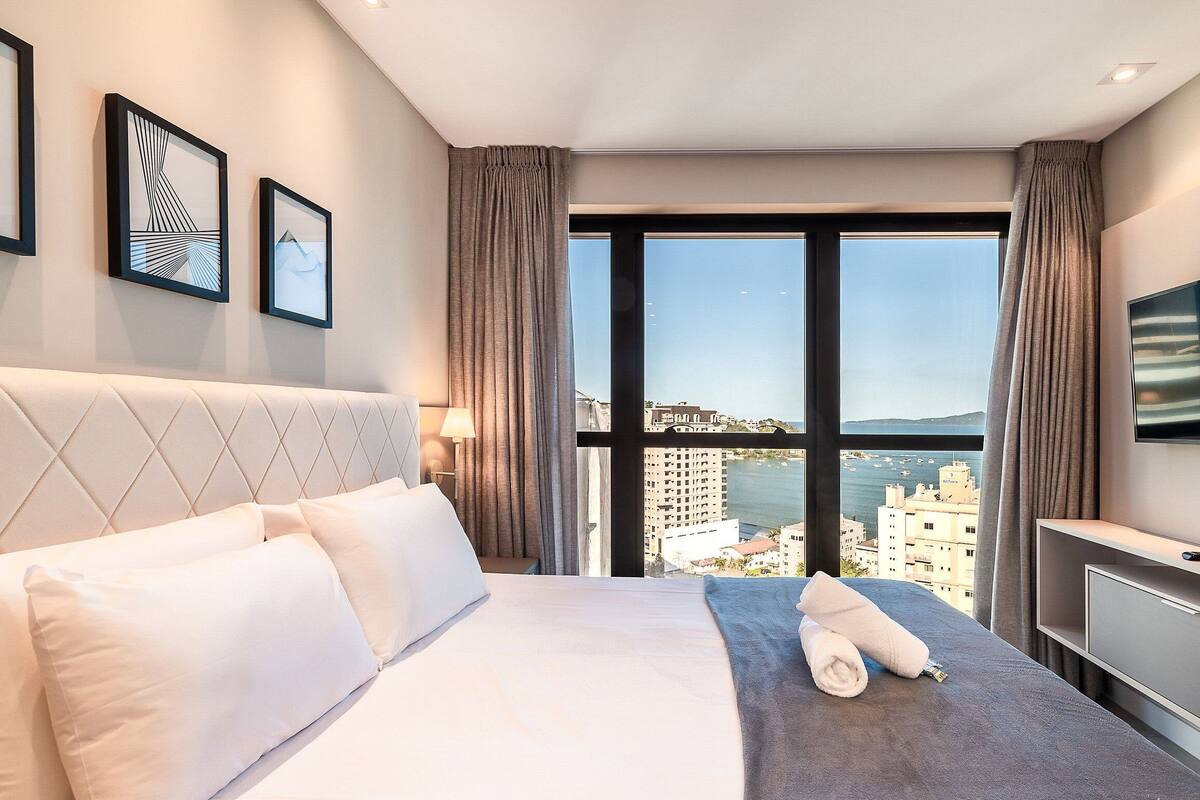 A hotel room with a large bed, a TV, a view of the sea, and framed artwork on the wall.