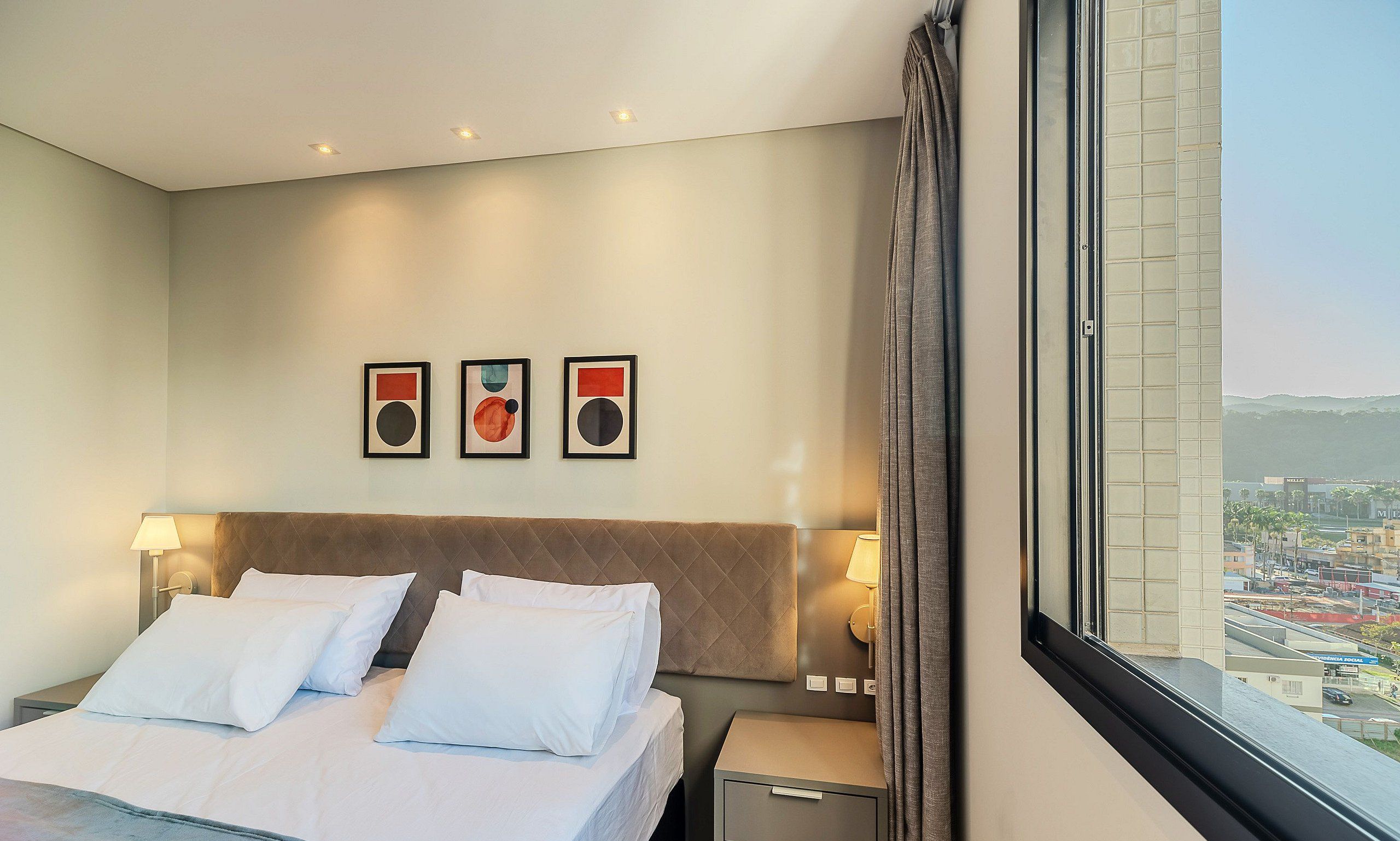 Ein Hotelzimmer mit Bett, Nachttischlampen, einem Nachttisch und einem großen Fenster mit Blick auf die Stadt.