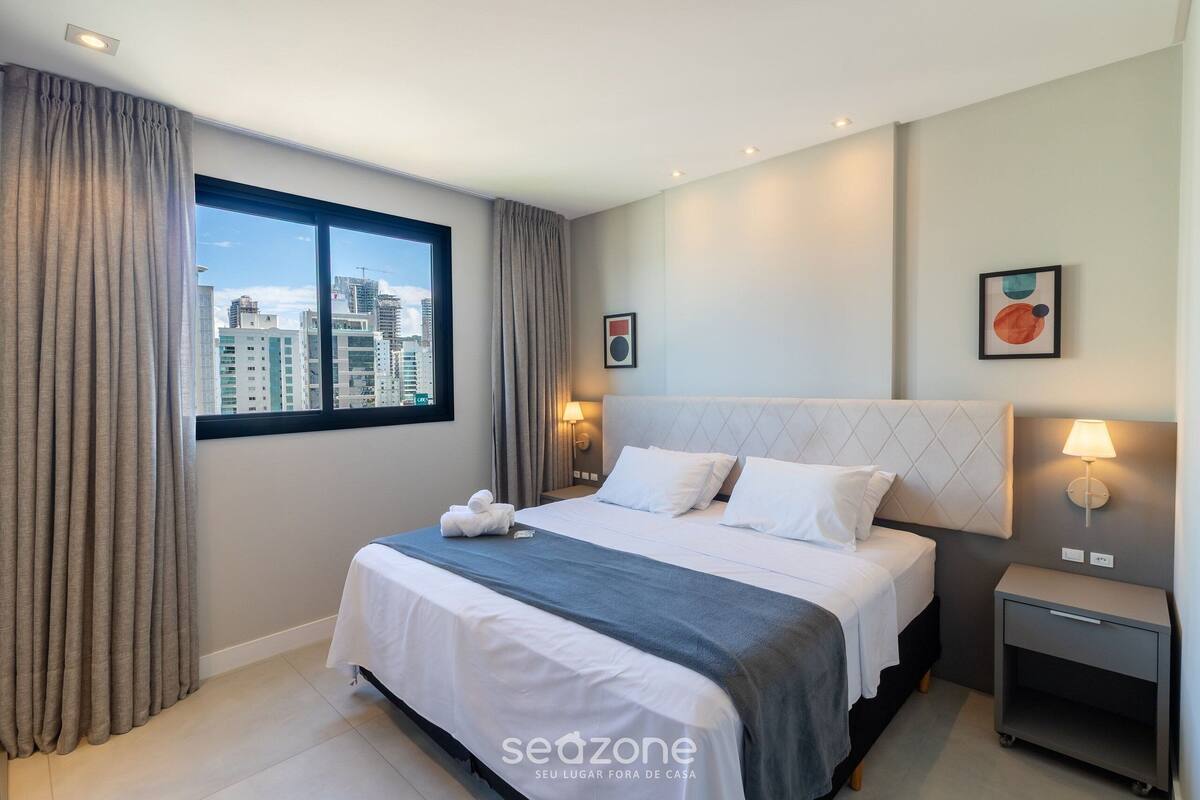 A modern hotel room with a large bed, a nightstand, a lamp, and a city view.