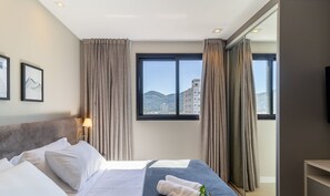 A hotel room with a bed, a nightstand, a lamp, a window with curtains, and a view of a cityscape. - MHF II Apts Variados em Prédio com Vista para o Mar em Itapema (Itapema)