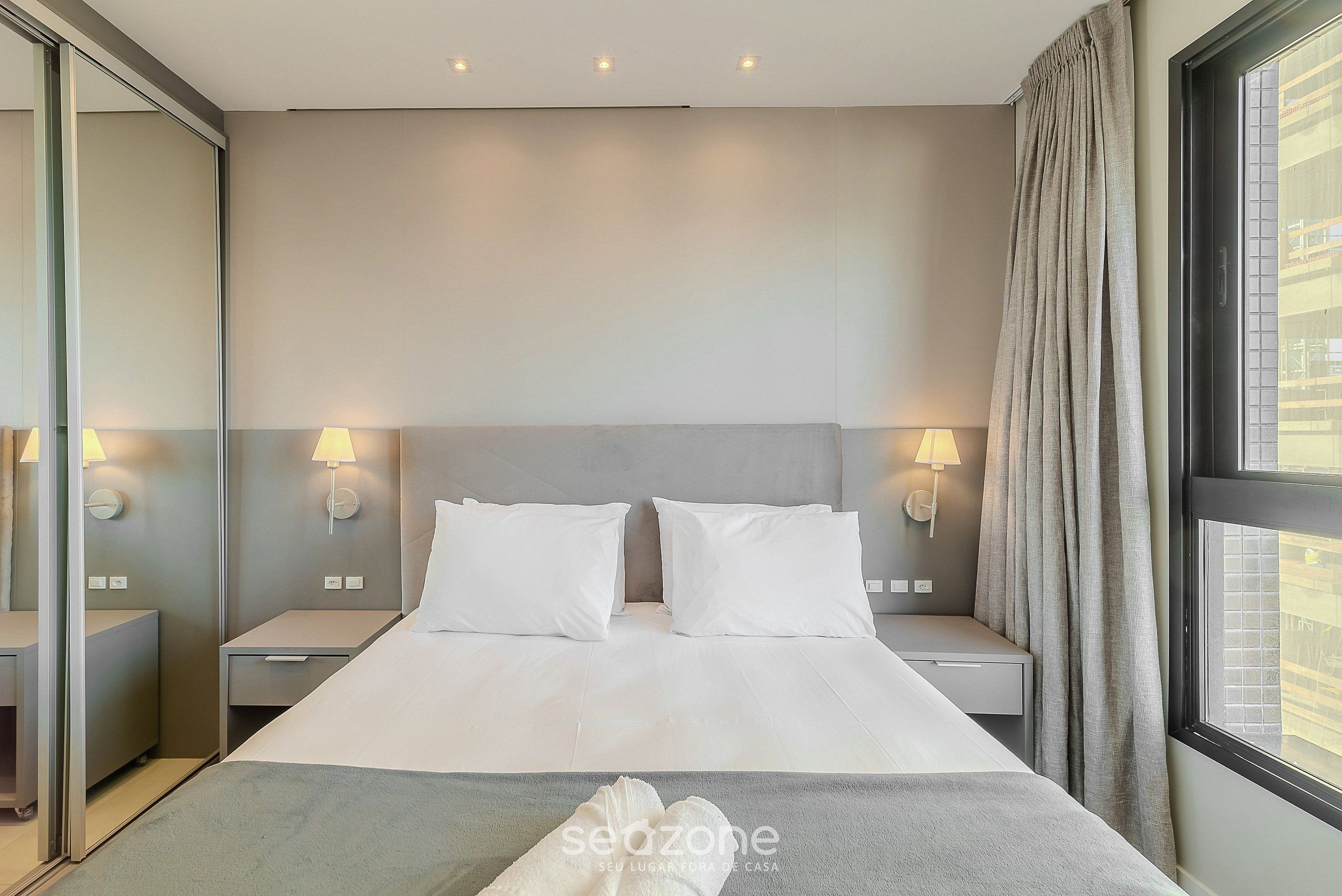 A hotel room with a large bed, two bedside tables, two wall-mounted lamps, a window with curtains, and a view of buildings outside.