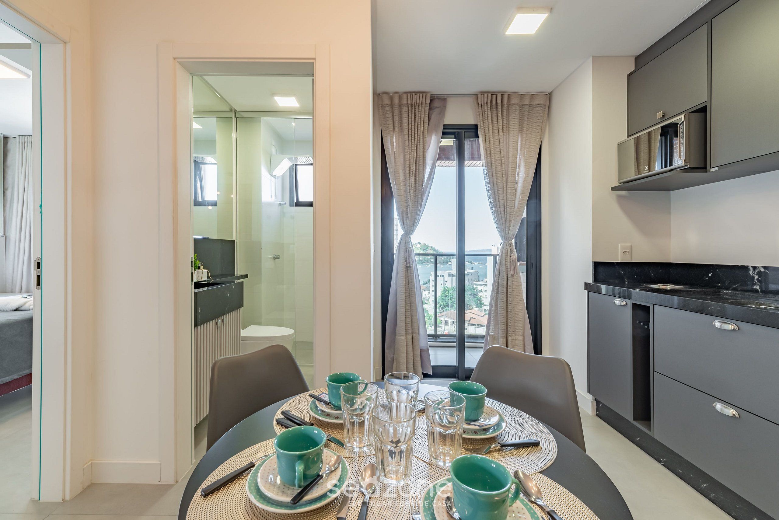 Una cocina moderna con armarios oscuros, una mesa de comedor con platos verdes y vasos transparentes, y vistas de la ciudad a través de amplios ventanales.