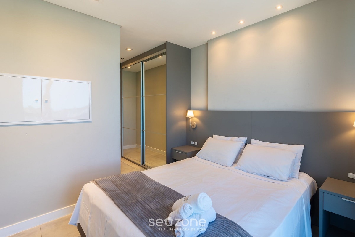 A modern hotel room with a large bed, a bedside table, a nightstand, and a wall-mounted lamp.