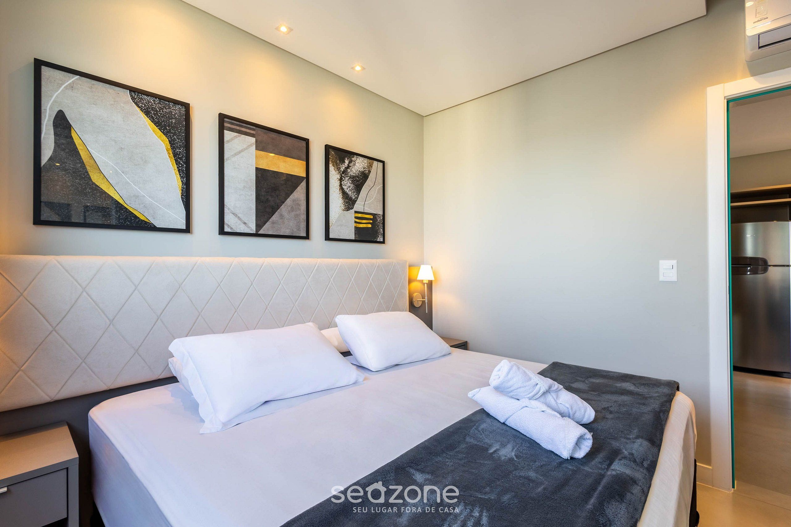 A hotel room with a bed, white pillows, a dark blanket, and three abstract paintings on the wall.