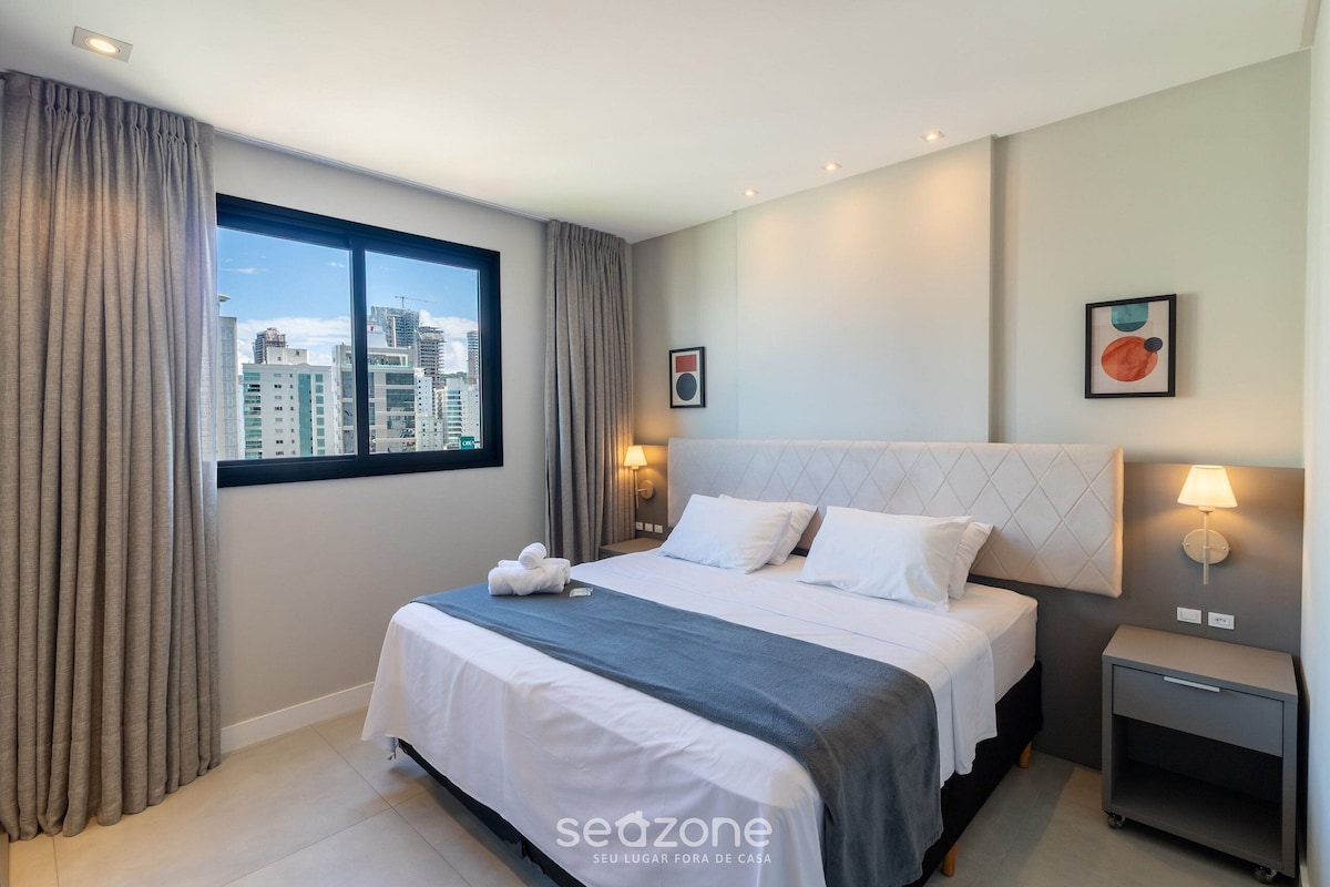 A modern hotel room with a large bed, a nightstand, a lamp, and a city view.