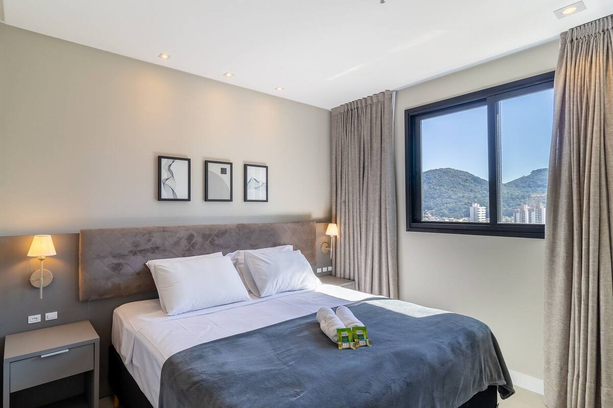 A hotel room with a bed, bedside lamps, a window with curtains, and artwork on the wall.