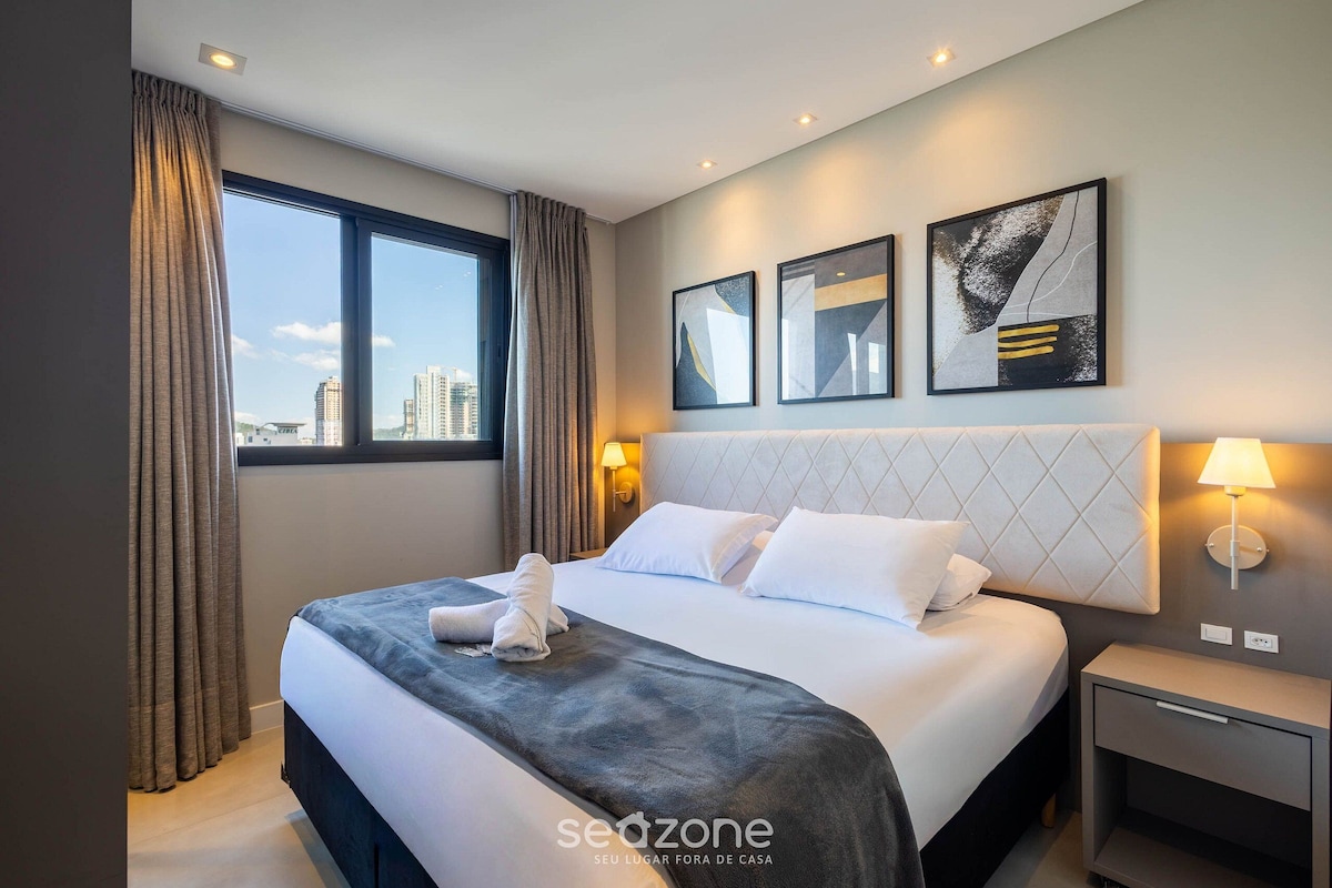 A hotel room with a large bed, a nightstand, a wall-mounted lamp, and a city view through the window.