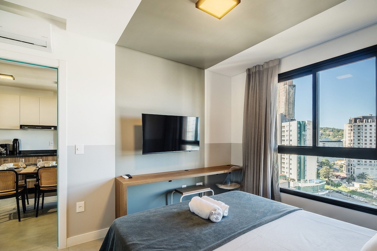 A hotel room with a bed, a TV mounted on the wall, a dining area with a table and chairs, and a large window with a city view.