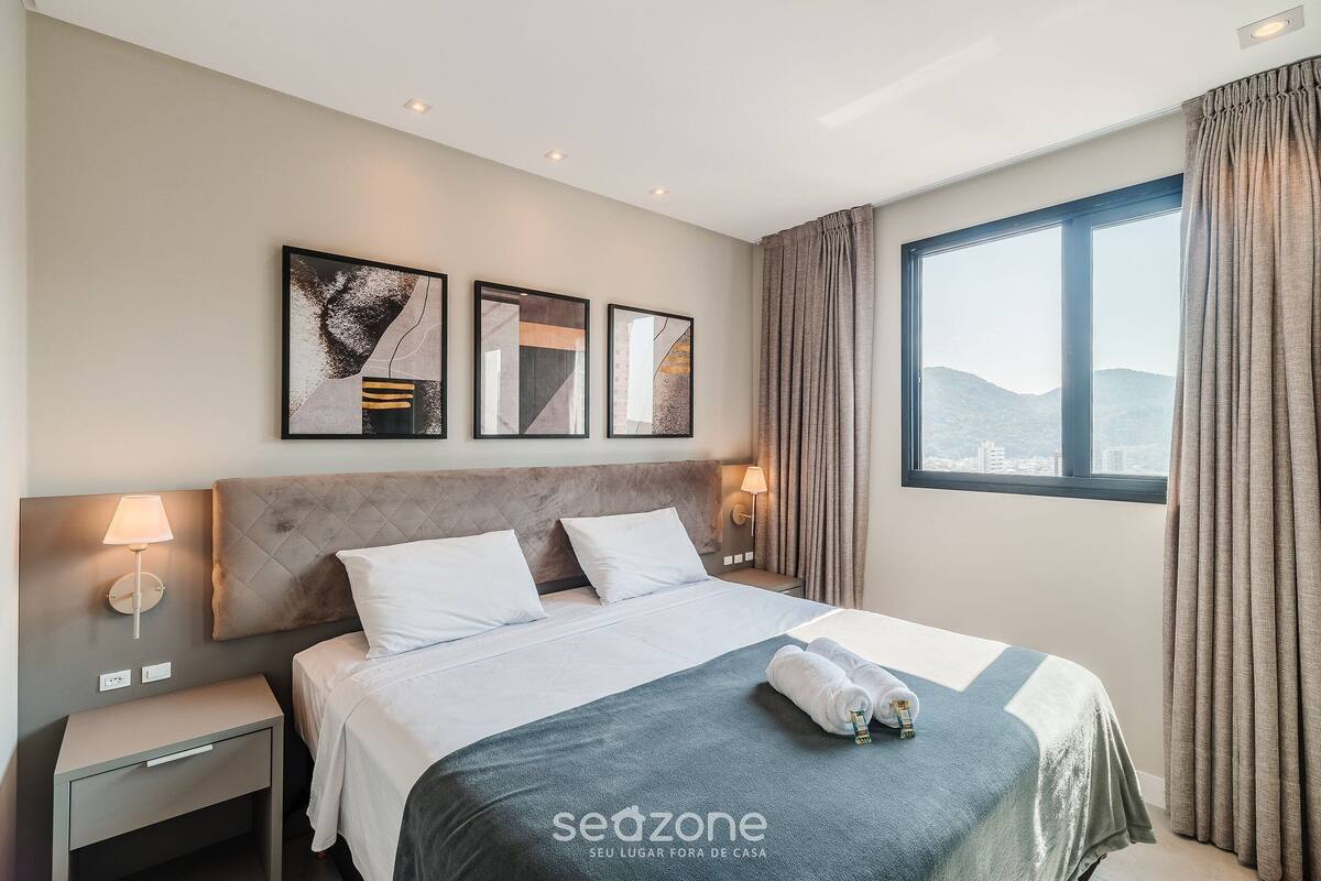 A hotel room with a bed, nightstand, wall art, and a window with curtains.