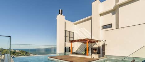 Une piscine à débordement située sur le toit, avec une terrasse en bois et des chaises longues, offrant une vue sur un paysage côtier.