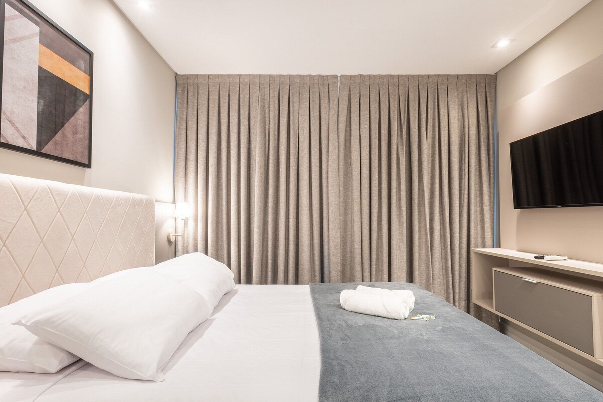 A hotel room with a bed, a flat-screen TV, a desk, and a window with curtains.