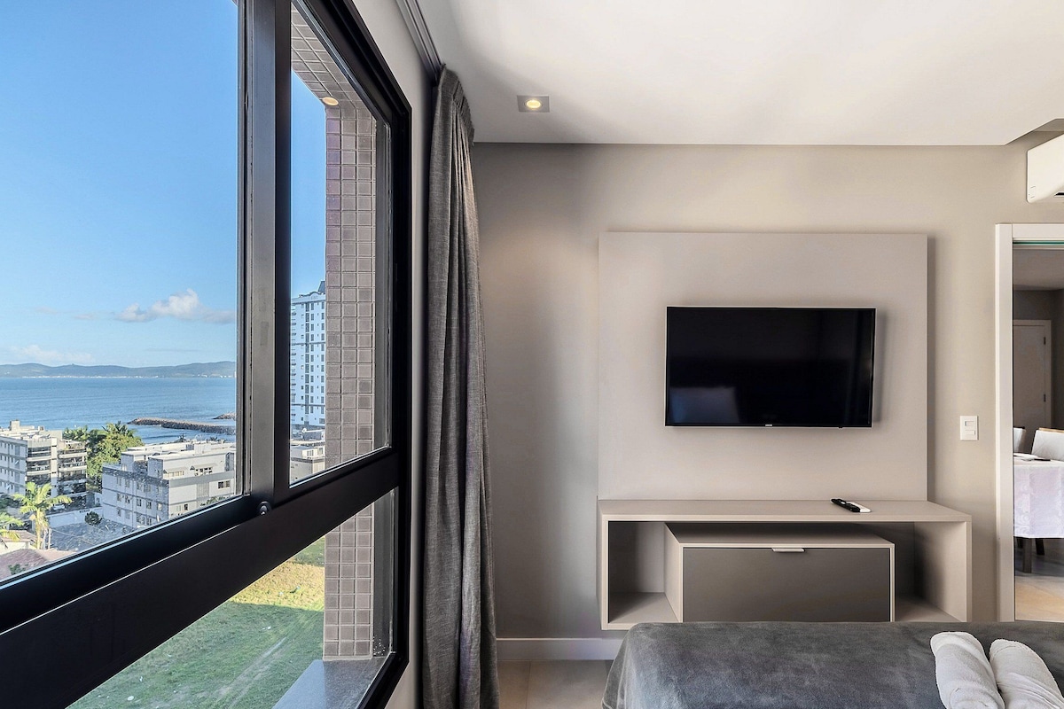 A modern hotel room with a large window, a flat-screen TV mounted on the wall, a small desk, and a bed with a view of the city and water.
