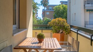 A balcony with a wooden table and chairs, a potted plant, and a view of buildings and trees.