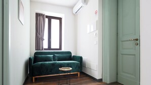 Una stanza con un divano verde, un tavolino rotondo, una finestra con tende, una porta e un condizionatore d'aria.