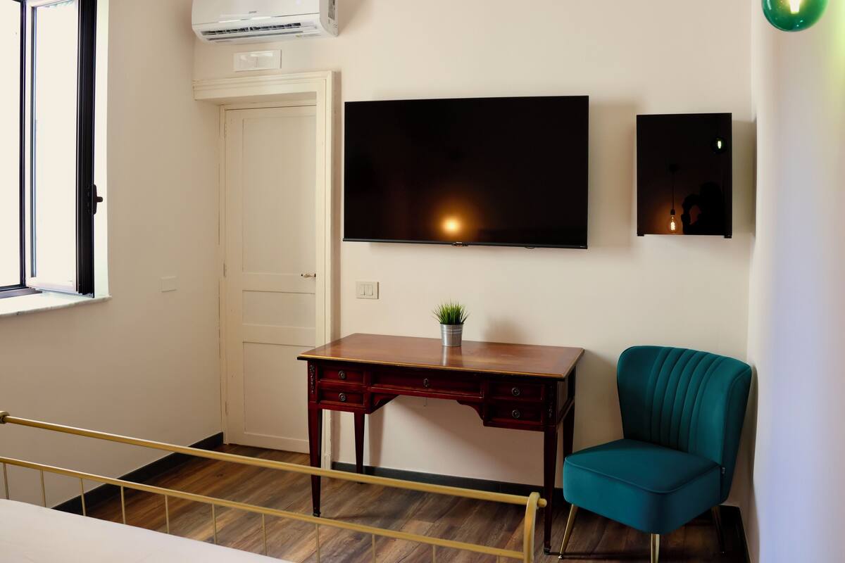 A room with a wooden desk, a teal chair, a flat-screen TV, and a small plant on the desk.