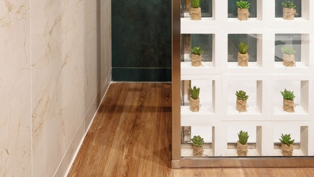 Un bagno con pareti in marmo, una parete d'accento verde scuro e un pavimento in legno. Si nota un mobiletto a scaffali bianco con piante in vaso.