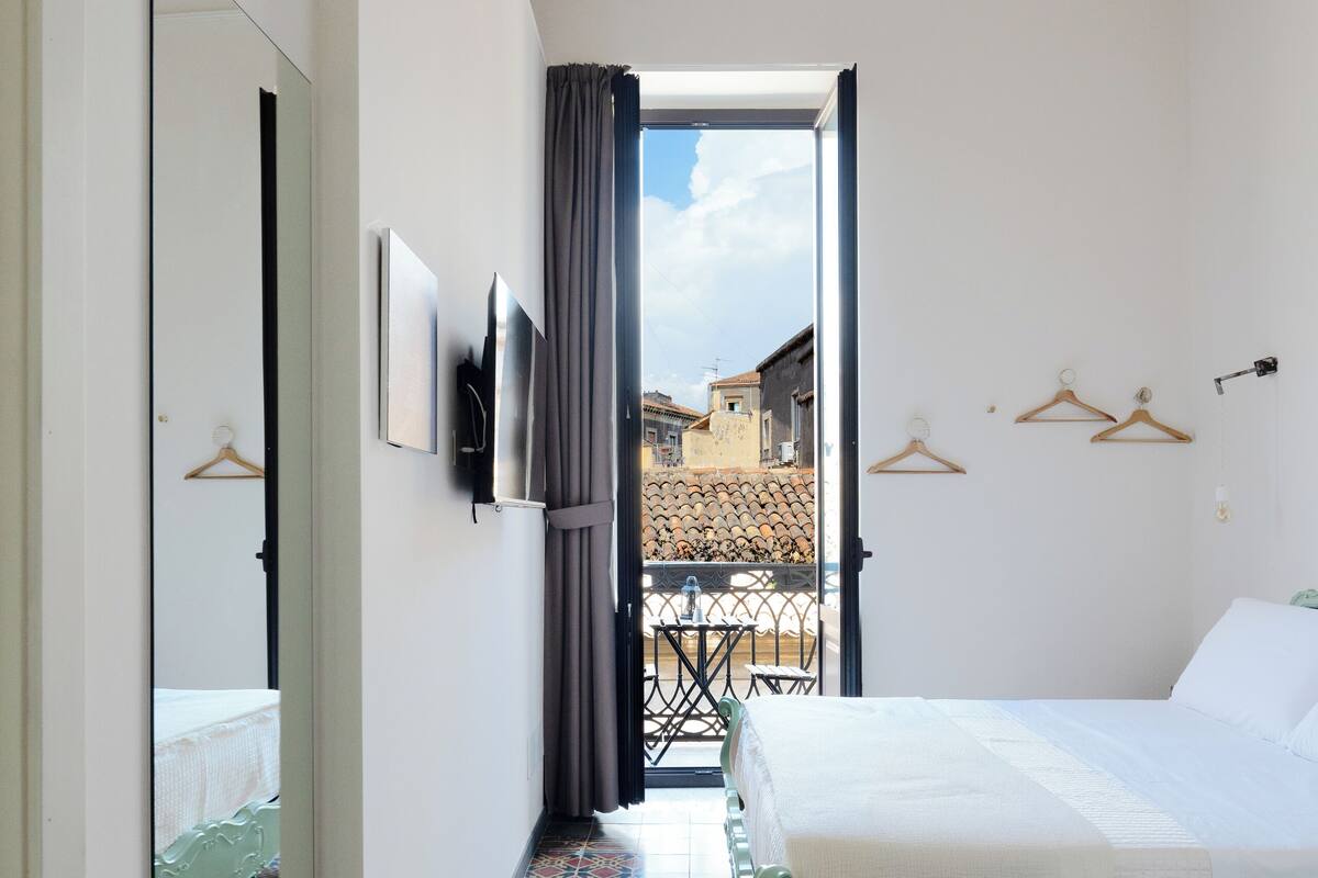 A hotel room with a bed, a mirror, a TV, and a view of the outside.