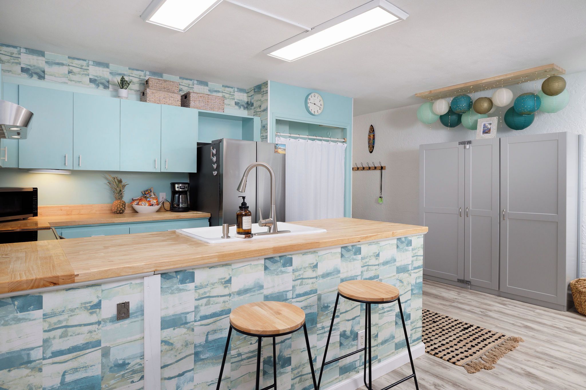 A modern kitchen with light blue cabinets, a wooden countertop, two stools, a refrigerator, and a clock on the wall.