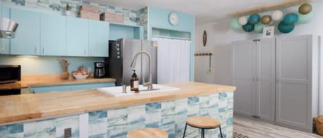 A modern kitchen with light blue cabinets, a wooden countertop, two stools, a refrigerator, and a clock on the wall.
