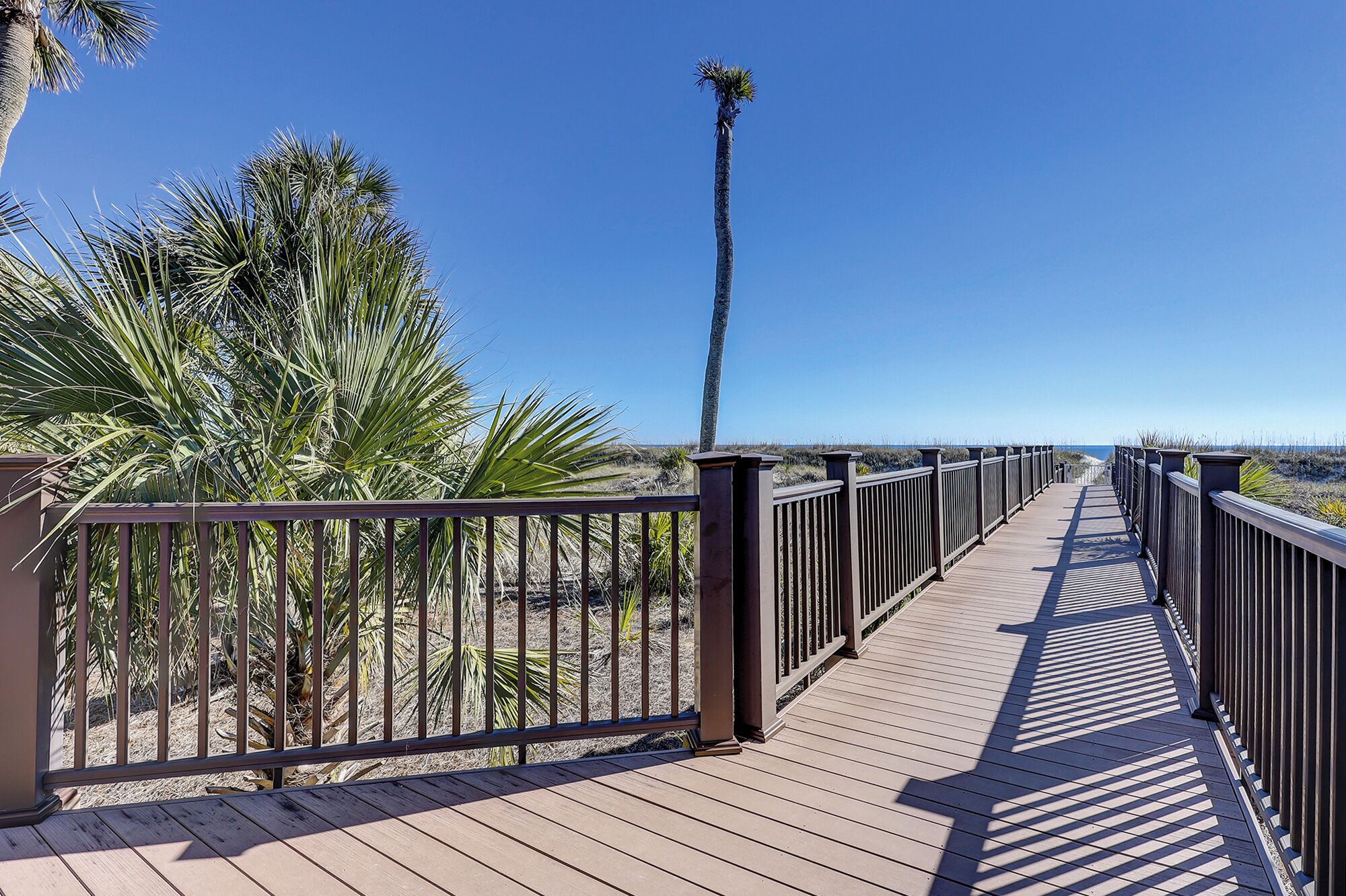 A wooden boardwalk with metal railings, leading to a beach with palm trees and a clear blue sky.