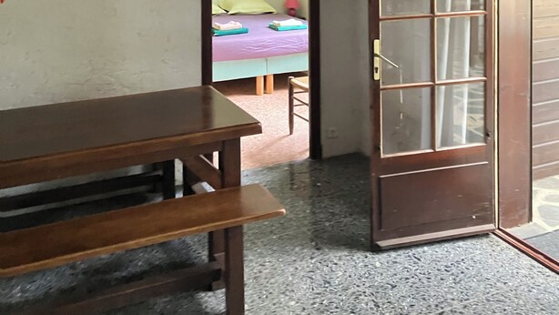 A room with a wooden table, a bench, a door with glass panels, and a bed with a green pillow and a pink lamp. - Gîte du Puy des Gardes (Meymac)